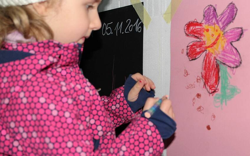 Ein Mädchen gestaltet eine bunte Blume auf Papier, das an der Wand eines Kern-Hauses hängt. 