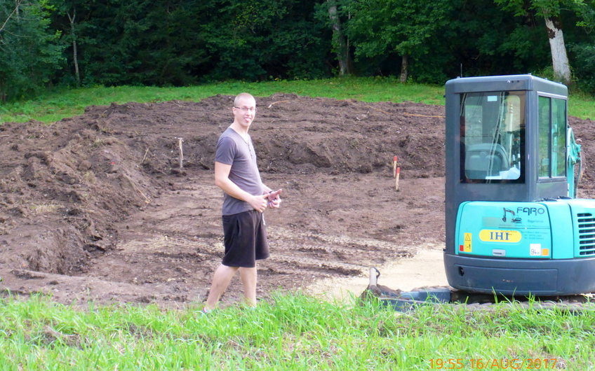 Baugrube mit abgetragenem Mutterboden und Mini-Bagger sowie Bauherr.
