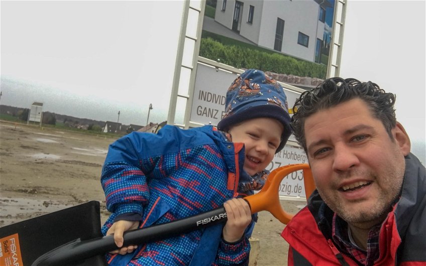 Bauherr mit Sohn und Schaufel vor Bauschild für Kern-Haus in Großpösna