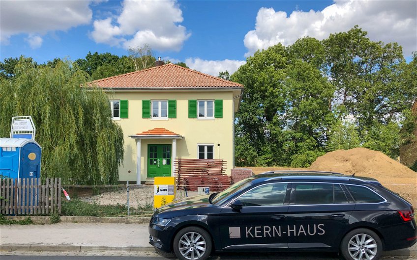 Kern-Haus Stadtvilla mit gelbem Putz, grünen Fensterläden, grüner Tür, rotem Dach und Kern-Haus Auto in Erdmannshain