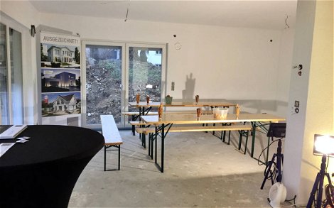 Verputzter Rohbau-Raum mit Bierbänken und Kern-Haus Info-Plakat.
